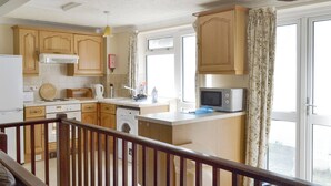 A kitchen with wooden cabinets, a microwave, a washing machine, and a dining area with a table and chairs. - Pirates Cove - UK44493 (Hope Cove, near Kingsbridge)