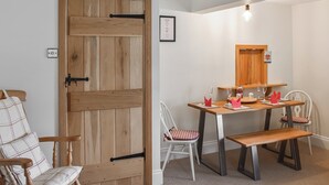 Un coin repas avec une table en bois dressée pour quatre personnes, un banc et deux chaises blanches dotées de coussins rouges. On aperçoit une porte en bois avec une poignée noire, et un petit tableau encadré est accroché au mur.