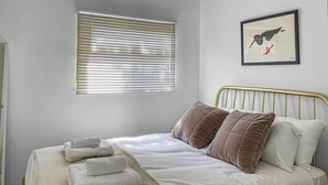 A neatly made bed with pillows and a blanket, a window with blinds, and a framed picture on the wall. - 2 bedroom accommodation in Cromer (Cromer)