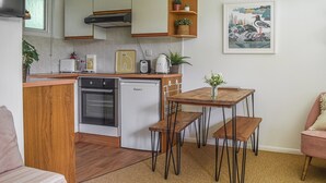 A compact kitchen with a dishwasher, a small refrigerator, a toaster, and a kettle. There is a wooden table with two benches and a small potted plant on it. - 2 bedroom accommodation in Cromer (Cromer)