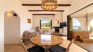 Een ruime eethoek met een ronde houten tafel en rieten stoelen, een grote geweven lamp en uitzicht op de omgeving door een groot raam.