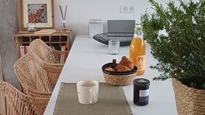 Uma sala de jantar com uma mesa branca, cadeiras de vime, uma planta em vaso, uma garrafa de suco, uma tigela de pão e uma caneca.