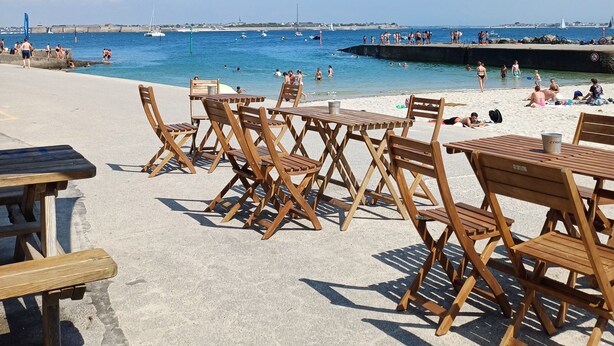 Um café à beira-mar com mesas e cadeiras de madeira, com vista para um mar azul cristalino e barcos, com pessoas.