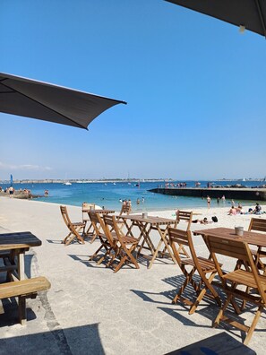 A beachside café with wooden tables and chairs, overlooking a clear blue sea with boats and people.