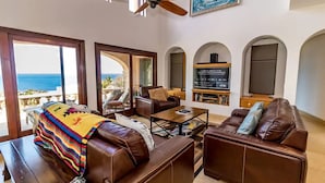 A spacious living area with a view of the ocean, leather furniture, a television, and a bookshelf.
