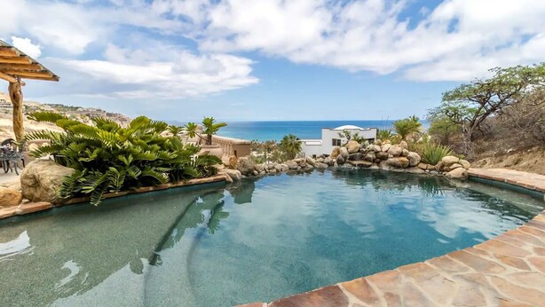 A clear blue pool with a view of the ocean, surrounded by rocks and tropical plants.