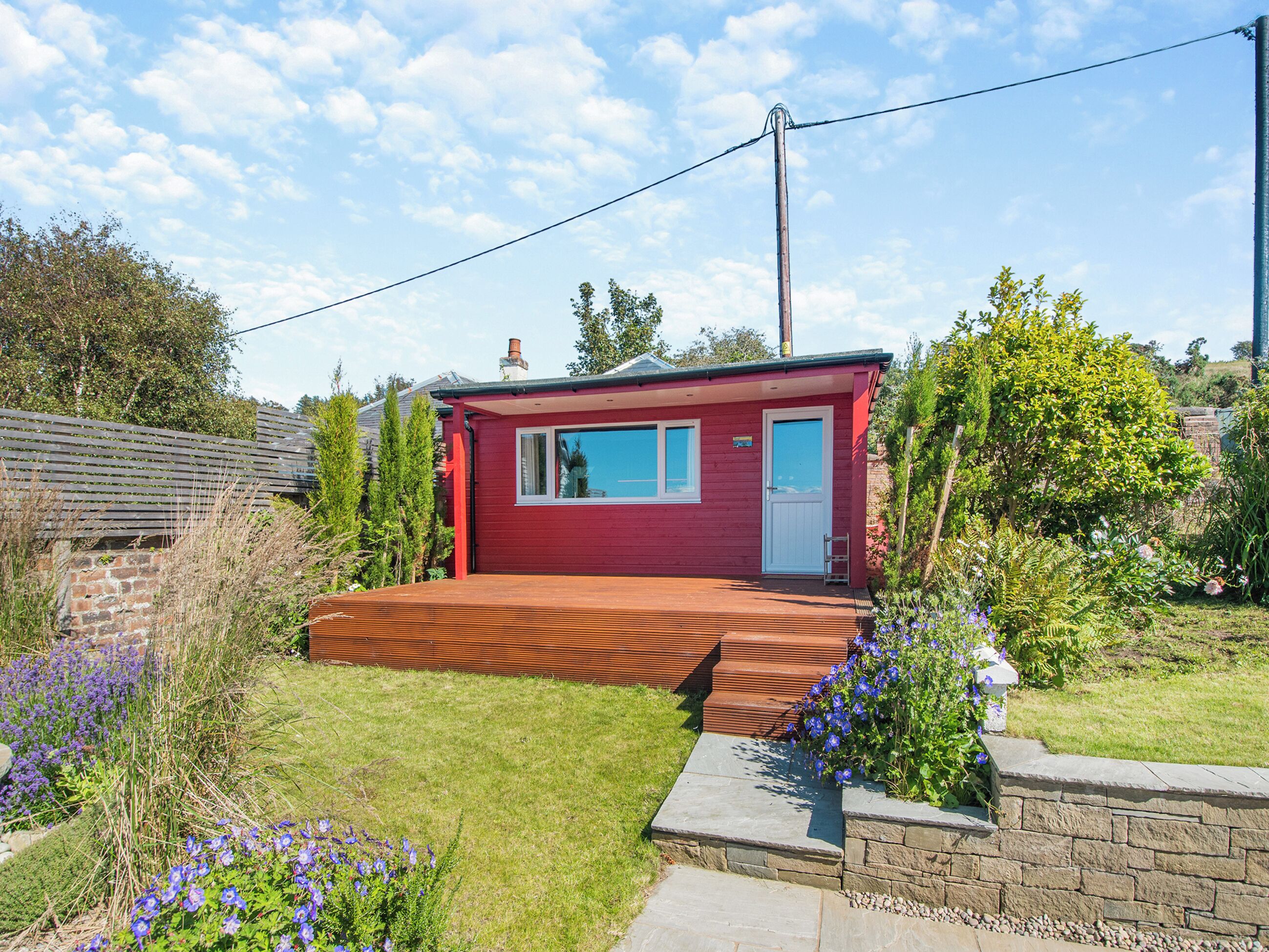 A red single-story house with a front porch, a small garden with purple flowers, and a clear blue sky.