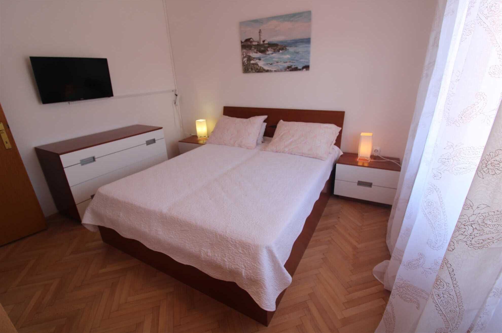 A bedroom with a wooden bed, two bedside tables with lamps, a flat-screen TV on the wall, and a painting above the bed.
