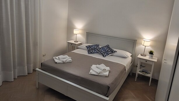 A modern hotel room with a single bed, two bedside tables with lamps, a ceiling light, and a window with curtains.