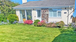 A single-story house with a tiled roof, a chimney, and a well-maintained lawn with various flowering plants. - A great, three-bedroom bungalow, with front and back gardens. (Warbstow, near Crackington Haven)