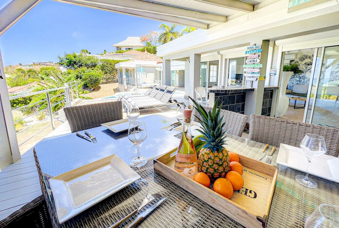 Ein Essbereich im Freien mit Korbmöbeln, einem Tisch gedeckt mit Tellern, Gläsern und einer Weinflasche. Ein Holzkasten mit Orangen und einer Ananas steht daneben.