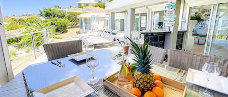 An outdoor dining area with wicker furniture, a table set with plates, glasses, and a bottle of wine. There is a wooden crate with oranges and a pineapple.