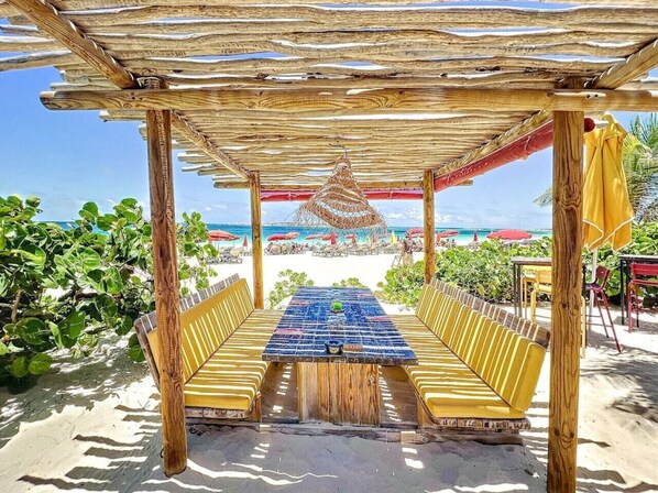 Un espace extérieur aménagé au bord de la plage, avec une pergola en bois, une table et des chaises, et une vue sur la plage et l’océan.
