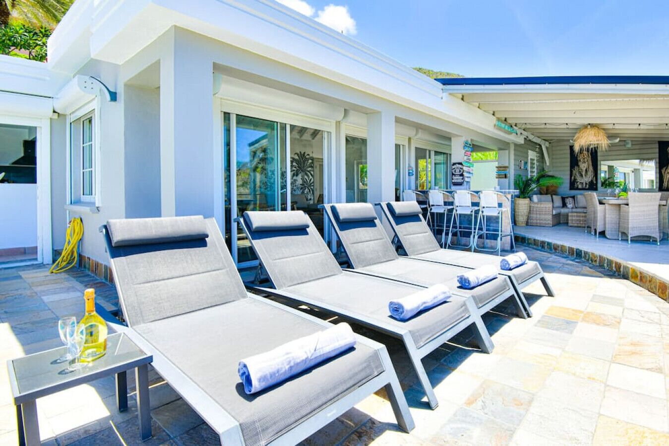 A poolside area with sun loungers, a table with a bottle, and a covered seating area with chairs and a table.