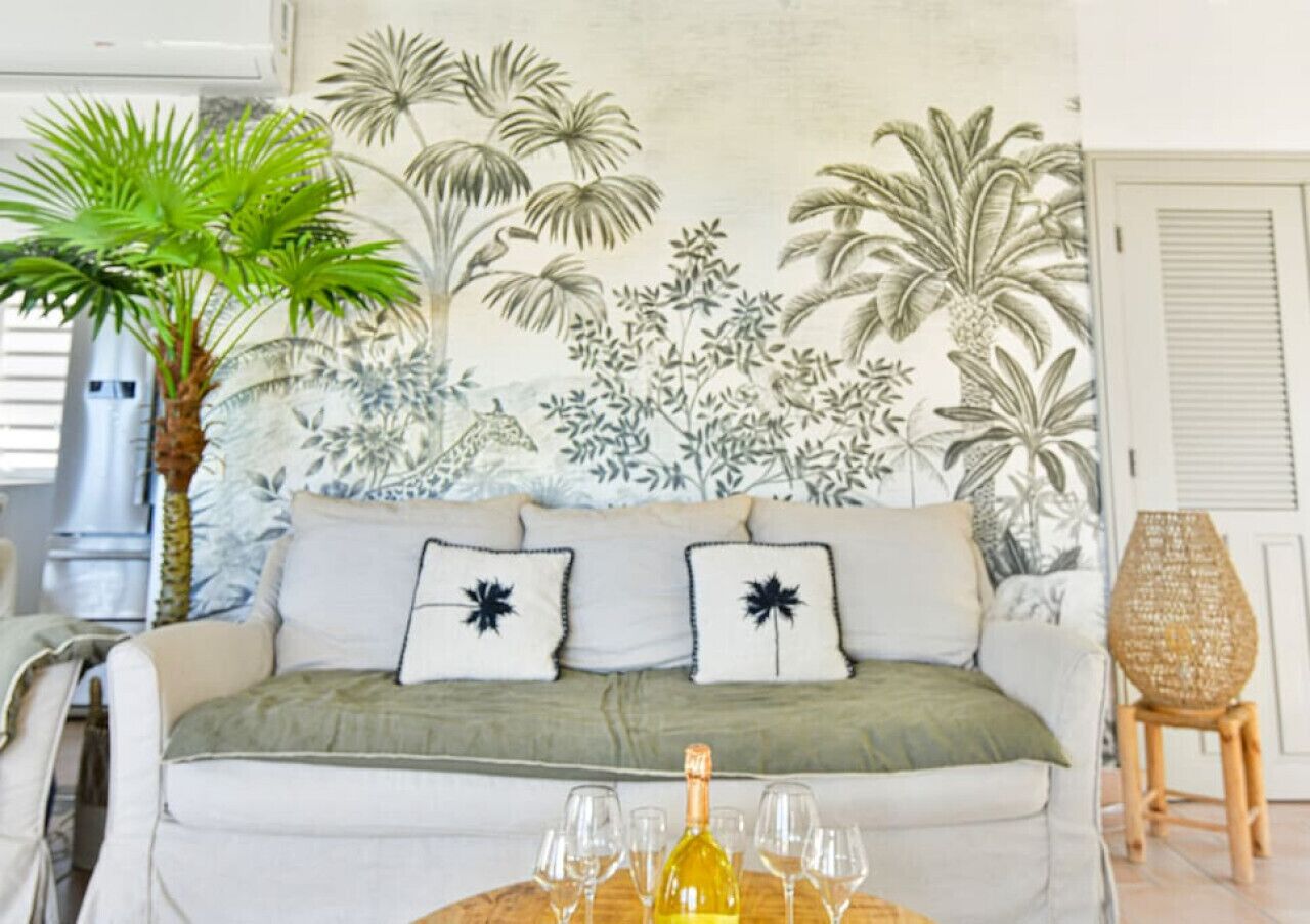 A living room with a sofa, two decorative pillows, a small table with glasses and a bottle, and a large wall mural of tropical plants.