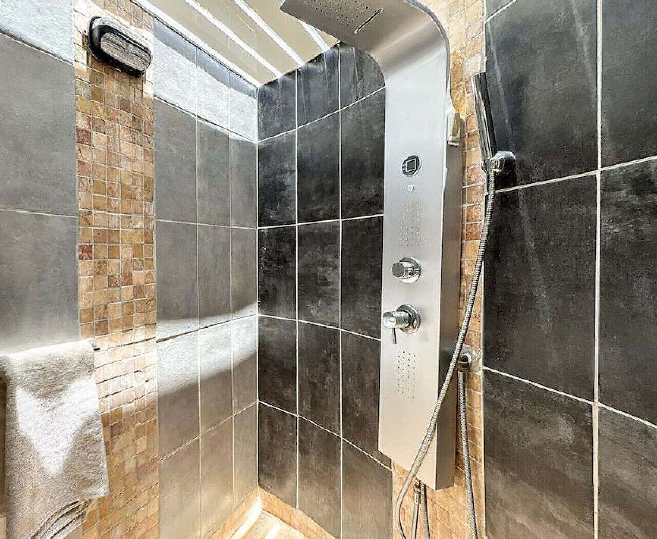 Une douche moderne dotée d’un pommeau de douche à main, d’un distributeur de savon intégré et d’un mur carrelé avec un mélange de carreaux de grande et de petite taille.