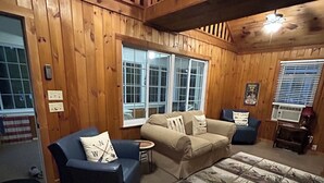 Ein gemütliches Blockhaus-Interieur mit Holzwänden, einem großen Fenster, einem blauen Sofa, einem beigen Sofa und einem gemusterten Teppich.