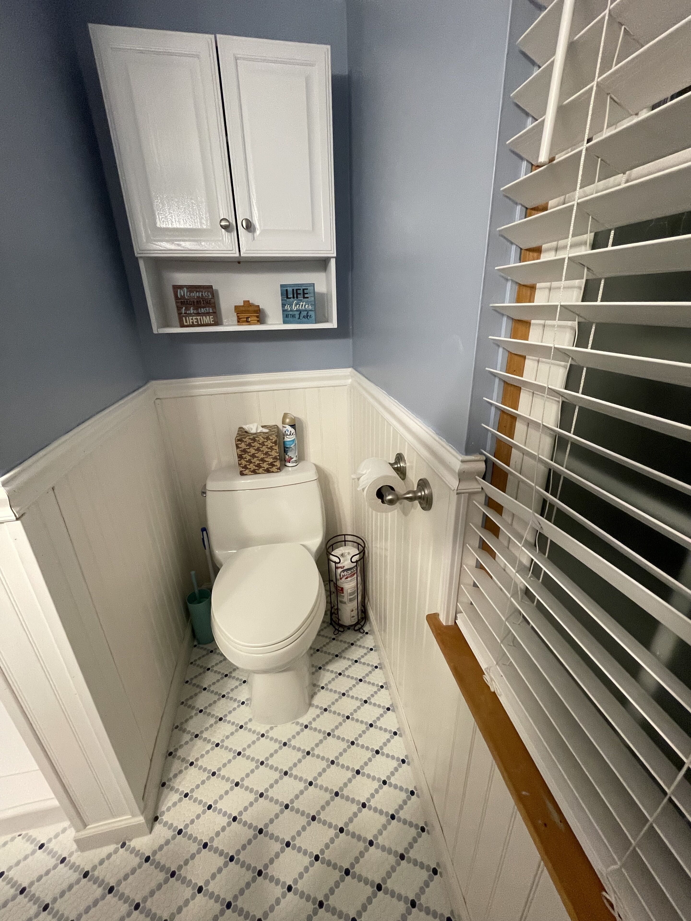 A bathroom with a toilet, a trash can, a cabinet above the toilet, and a window with blinds.