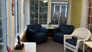 Ein Zimmer mit zwei dunkelblauen Stühlen, einem weißen Rattanstuhl, einem weißen Tisch und einem Fenster mit Blick auf Grünflächen.