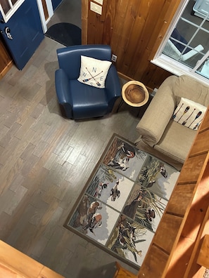 Ein Zimmer mit einem blauen Sessel, einem beigen Sofa, Holzboden und einem Glastisch mit Blumenmuster.
