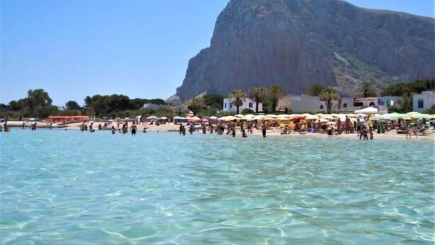 Una playa con agua cristalina, sombrillas y gente. Una gran montaña al fondo.