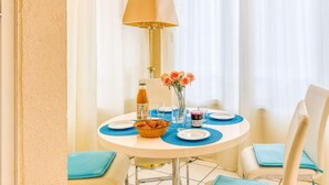 Una mesa de comedor redonda con individuales azules, un bol de croissants, una botella de jugo de naranja, un vaso de agua y un jarrón con rosas rosadas.