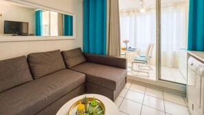 Una sala de estar moderna con un sofá seccional marrón, una pequeña mesa redonda de centro y un televisor sobre un mueble blanco. Hay una puerta corrediza de vidrio que da a un balcón con cortinas azules.