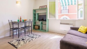Un espacio habitable pequeño con zona de comedor, una estantería verde, un ventanal con cortina a rayas y un sofá con un cojín amarillo.