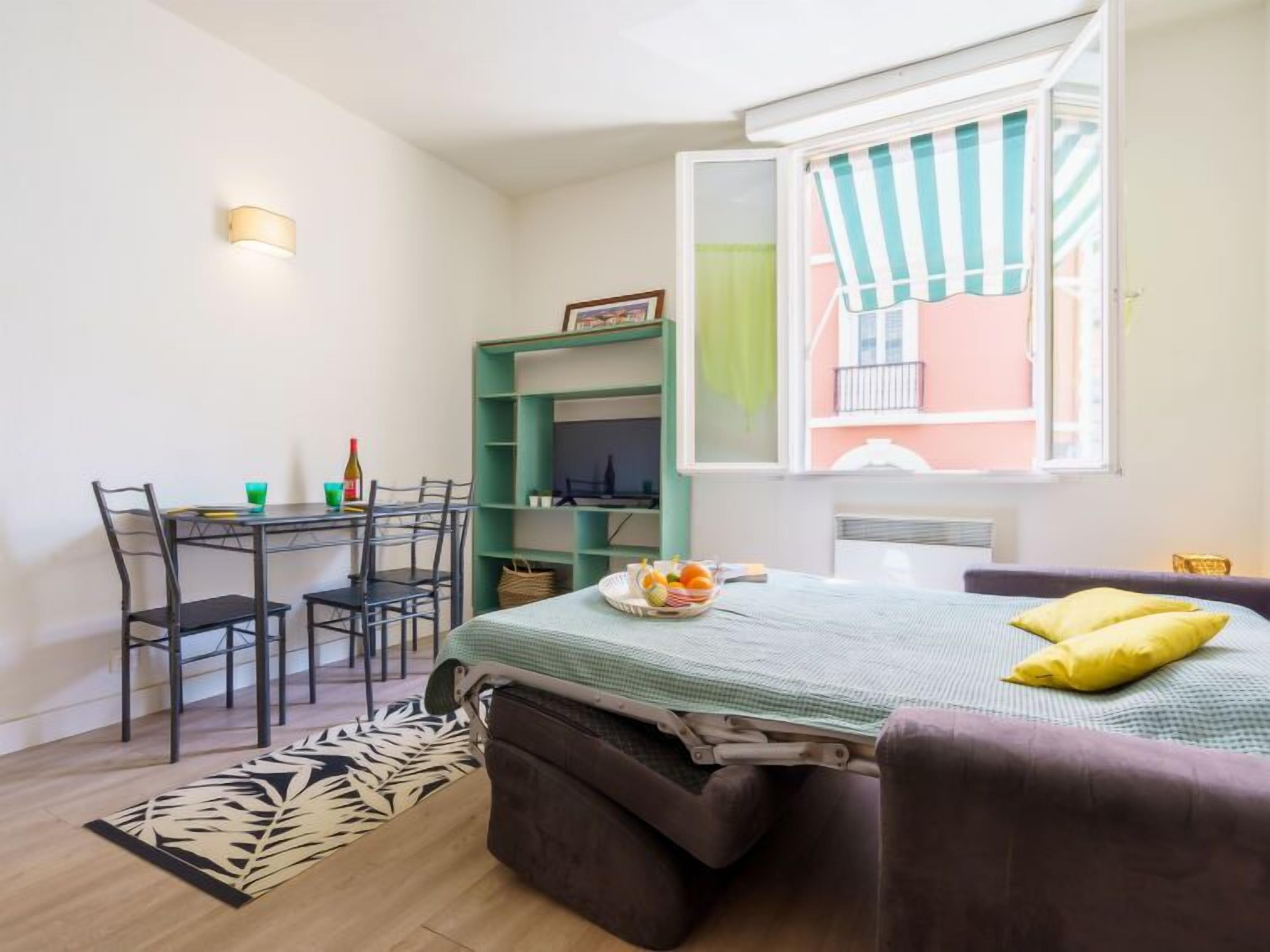 Ein kleines Zimmer mit einem Bett, einem Schreibtisch und einem Stuhl. Ein Fenster mit einem gestreiften Vorhang, ein kleiner Balkon und eine Wandleuchte sind vorhanden.