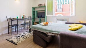 Habitación compacta con cama, escritorio y silla. Cuenta con una ventana con cortina a rayas, un balcón pequeño y una lámpara fijada a la pared.