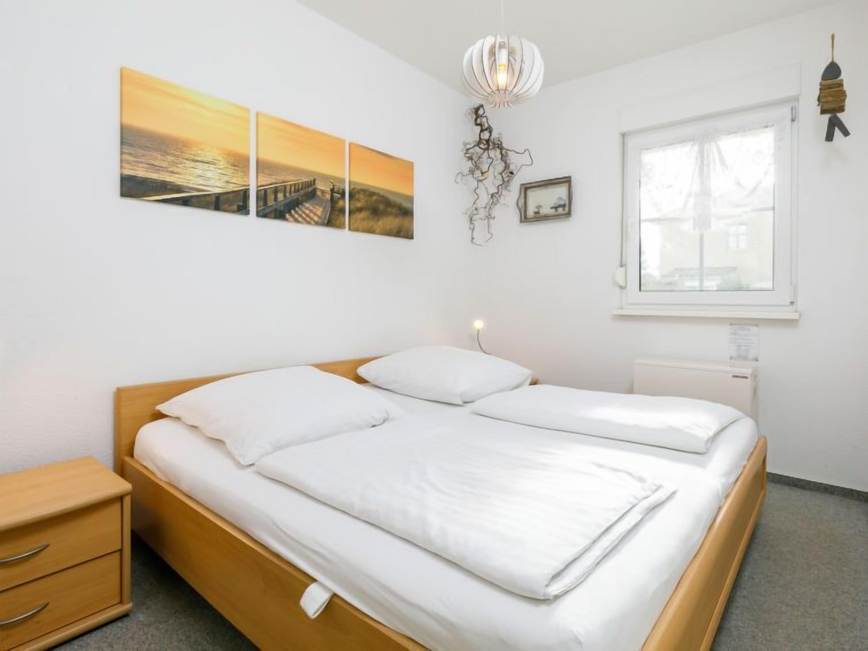 A bedroom with a wooden bed, white bedding, a nightstand, a bedside lamp, a window, and wall art.