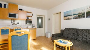 A compact living space with a kitchenette, a dining area with blue chairs, a grey sofa, and a small table with a magazine. - Apartment Berg by Interhome (Zinnowitz)