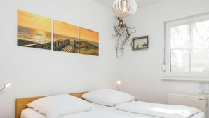 A neatly made bed with white linens in a room with a wooden headboard. There are two wall-mounted paintings depicting a sunset over a body of water and a bridge. A unique light fixture hangs from the ceiling, and a small framed picture is visible on the wall. - Apartment Berg by Interhome (Zinnowitz)