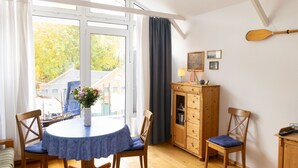 Ein gemütliches Zimmer mit einem runden Tisch, zwei Stühlen, einer Holzkommode und einem großen Fenster mit Vorhängen.
