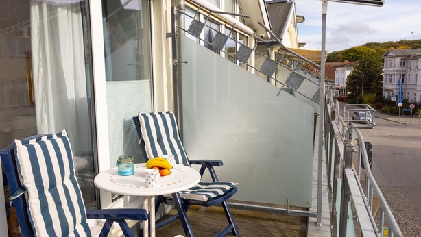 Ein Balkon mit zwei gestreiften Liegestühlen, einem kleinen Tisch mit Obstschale und einer Glastür, die zu einem Gebäude führt.