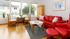 A bright living room with a red sofa, wooden chairs, a small table, and a view of a building through large windows. - Luise von Preußen (App. 205) by Interhome (Zinnowitz)