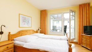 A bedroom with a wooden bed, a nightstand, a dresser, a television, and a large window with curtains. - Luise von Preußen (App. 205) by Interhome (Zinnowitz)
