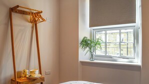 Un dormitorio con una cama, un tendedero de madera, una ventana con vistas a edificios y una planta en maceta en el alféizar.