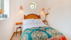 A bedroom with a wooden headboard, a teal floral bedspread, two pendant lights, a bedside table with a vase, and a window with a view of the outside.