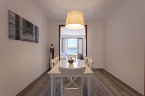 A dining area with a table and chairs, a pendant light, a painting on the wall, and a view of the sea through an open door.