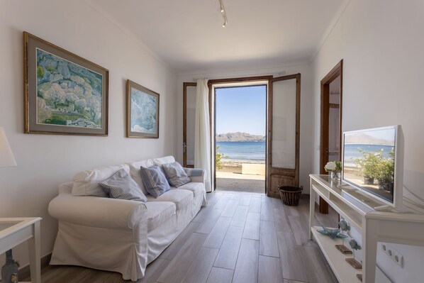 A room with a white sofa, a television on a white cabinet, and a view of the sea through an open door.