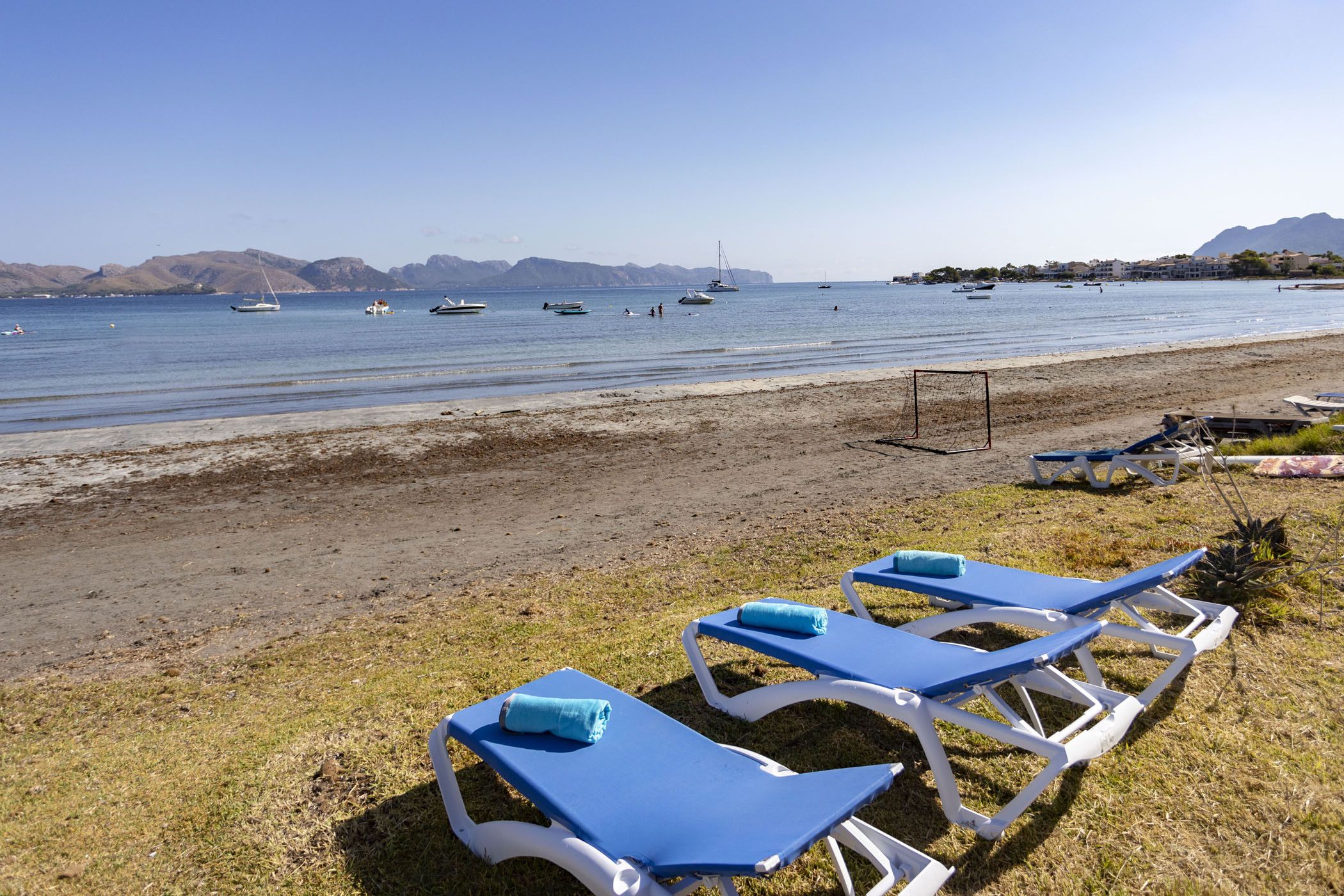 A beach with lounge chairs and a soccer goal, boats in the water, and mountains in the background.