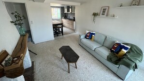 Una sala de estar con un sofá, una mesa de centro y un banco de madera. Se aprecia una cocina al fondo, con armarios y una encimera.
