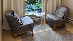 Une pièce comprenant deux fauteuils à motifs, une petite table ronde et une vue sur un jardin, visible à travers une fenêtre ornée de rideaux blancs.