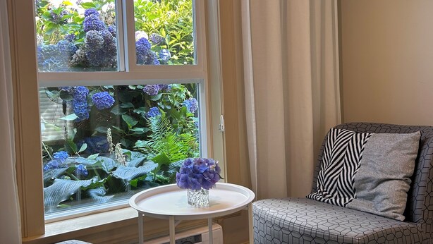 Une pièce avec une fenêtre donnant sur un jardin, une petite table ronde avec un vase et un fauteuil à motifs.