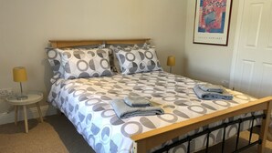 Een net opgemaakt bed met een geborduurd sprei, twee kussens en opgevouwen handdoeken ernaast. Er staat een klein nachtkastje met een lamp, en een ingelijst beeld op de muur.