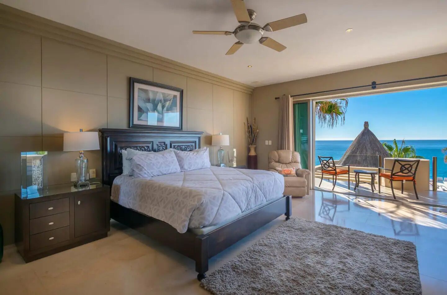 A spacious bedroom with a large bed, a ceiling fan, a nightstand, and a view of the ocean through a sliding glass door.