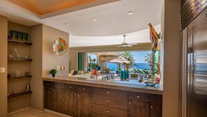 A kitchen with dark wooden cabinets, a granite countertop, and a view of an outdoor patio with a pool and lounge chairs. - Villa Alegria (Cabo San Lucas)