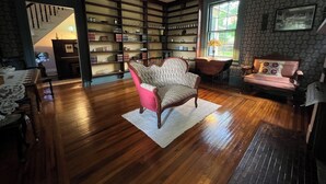 A spacious room with hardwood floors, a ceiling fan, a large bookshelf, and a cozy seating area with a chair and a sofa. - Highland Guesthouse (Meadville)