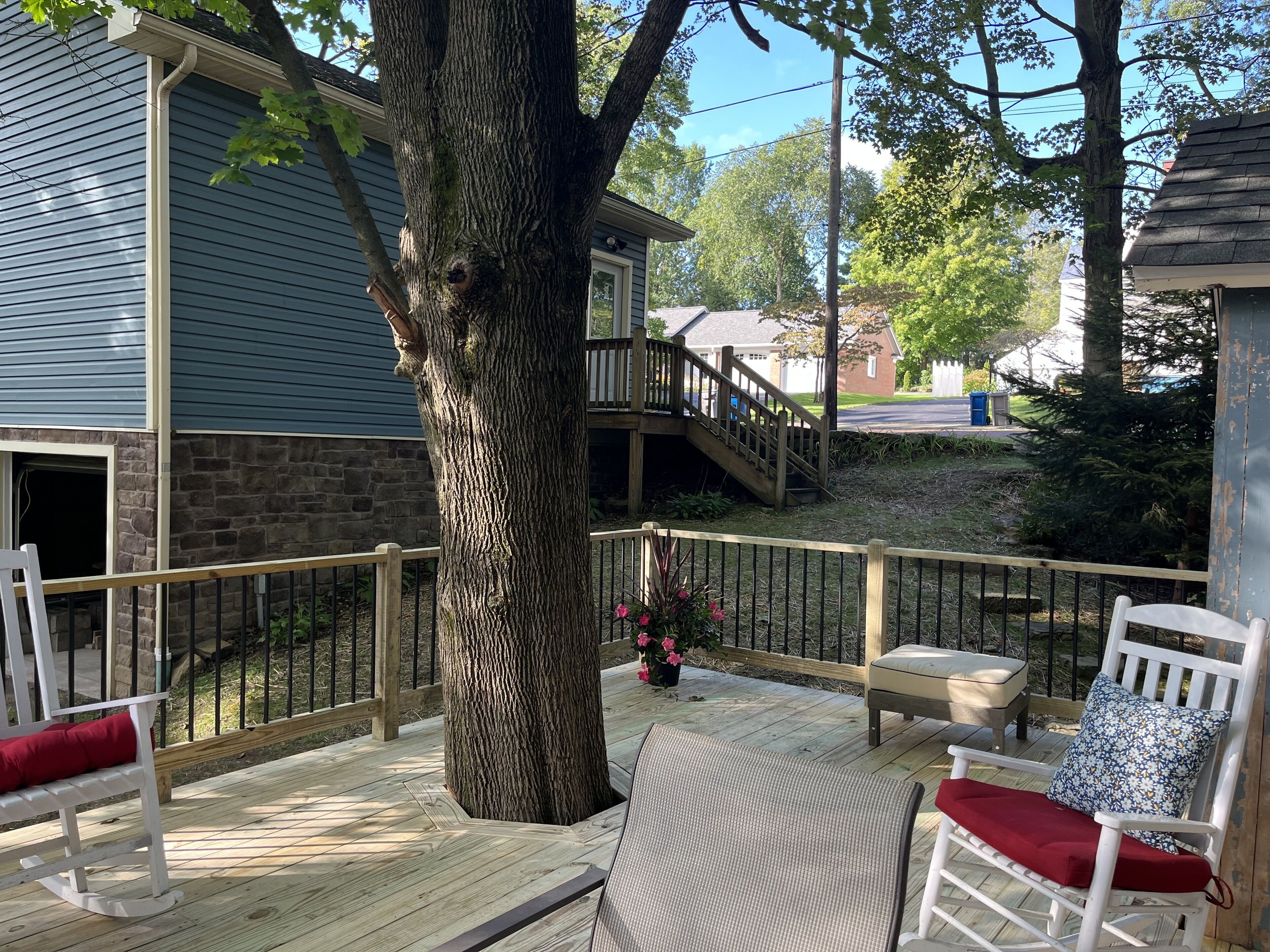 Une terrasse en bois avec un grand arbre, un mur en pierre et un escalier menant à un balcon situé au deuxième étage.